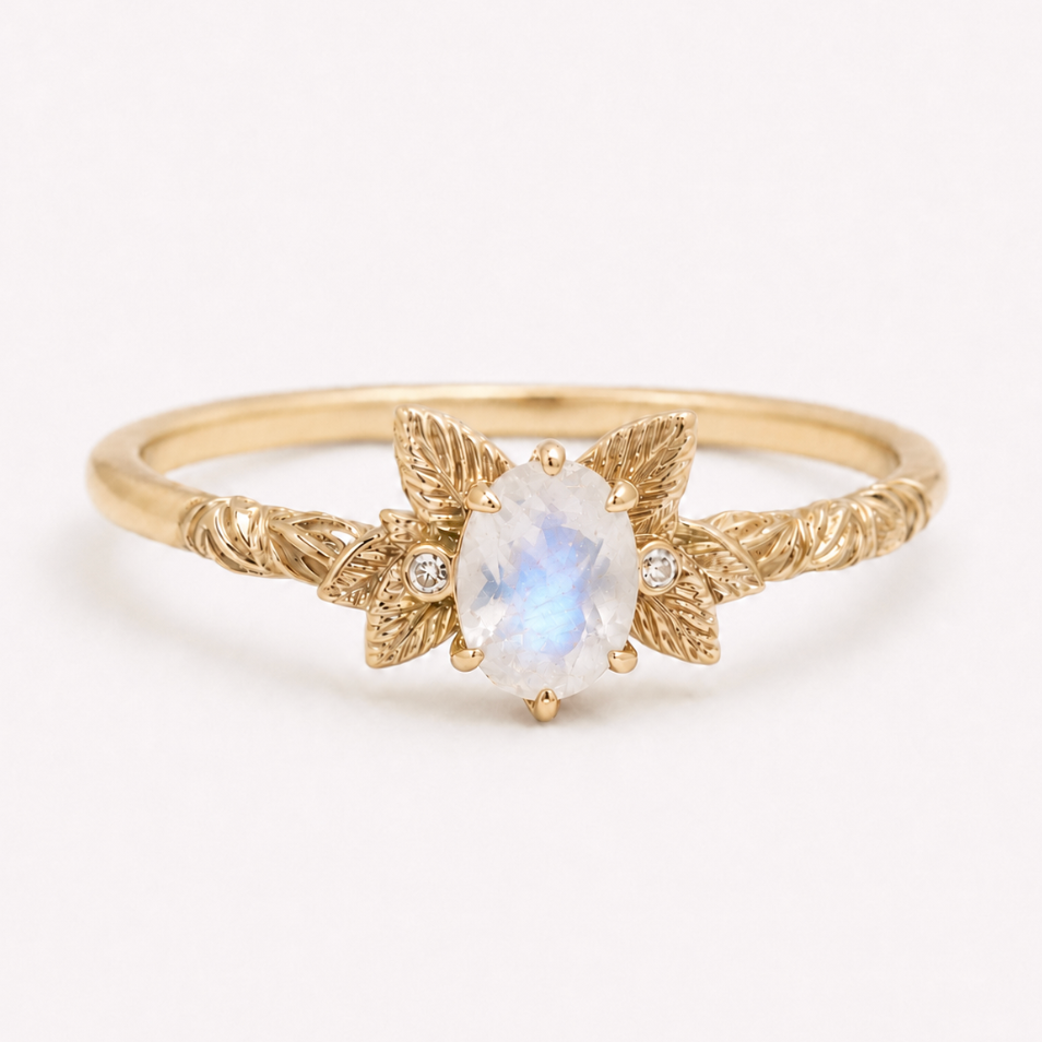 A gold ring featuring a central oval moonstone with a soft blue-green iridescent glow, flanked by two textured leaf motifs and small clear accent stones, set against a plain white background.