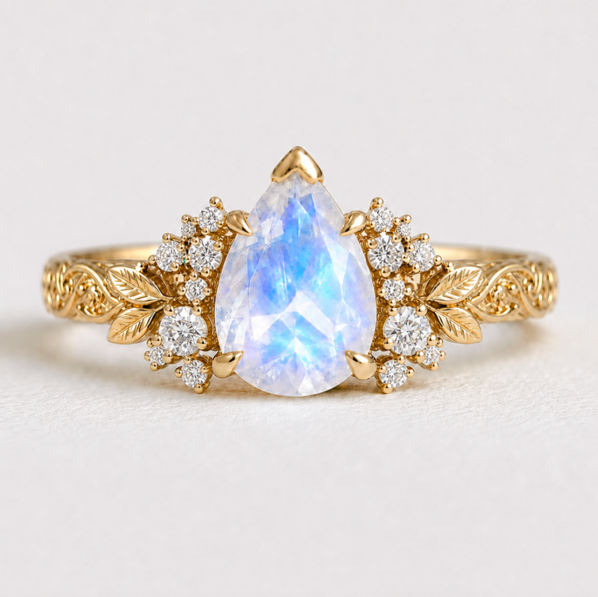 A gold ring featuring a pear-shaped moonstone centerpiece with a blue iridescent sheen, flanked by clusters of small clear stones and leaf-shaped gold details on a plain light background.