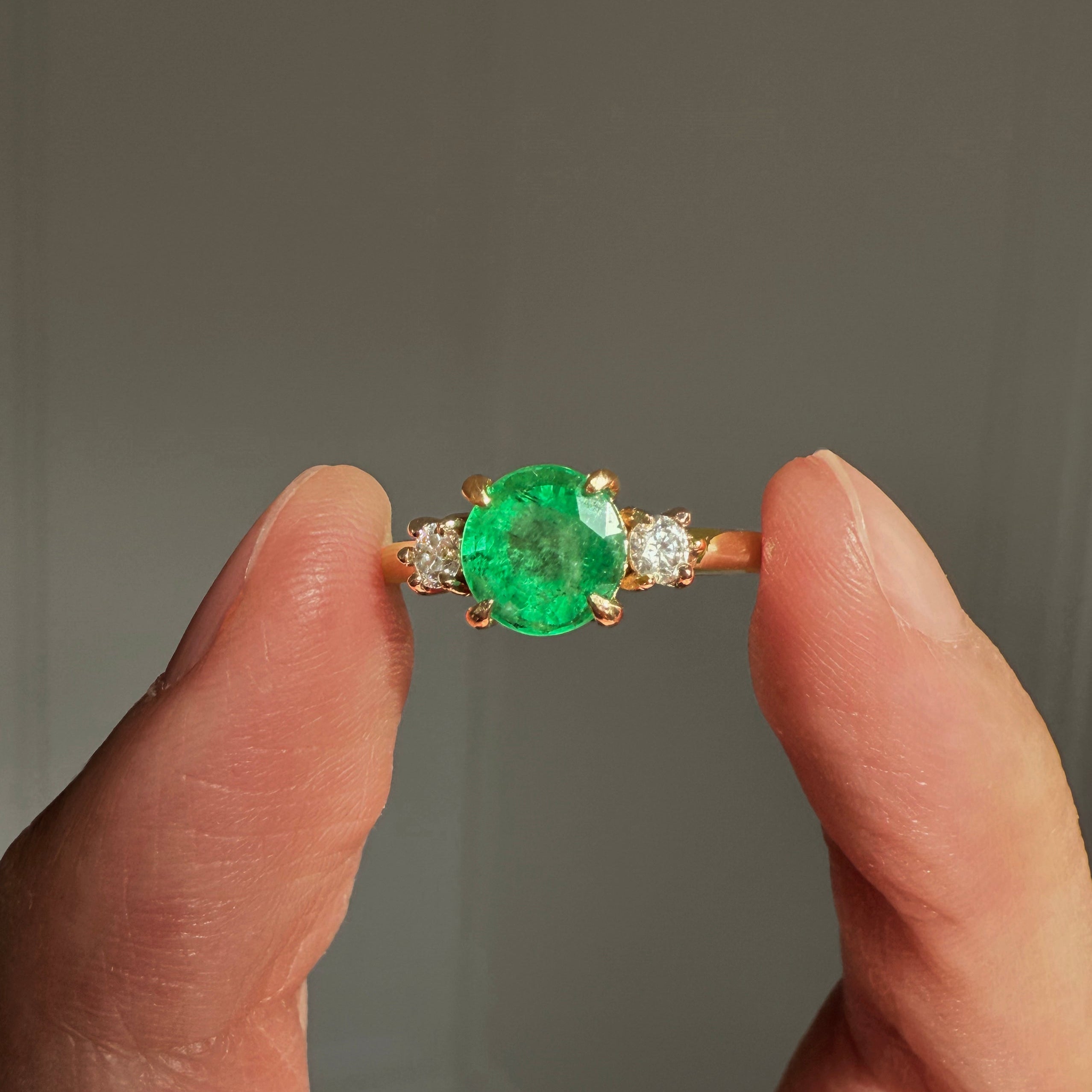 Gold ring with a green emerald held between two fingers against a neutral background