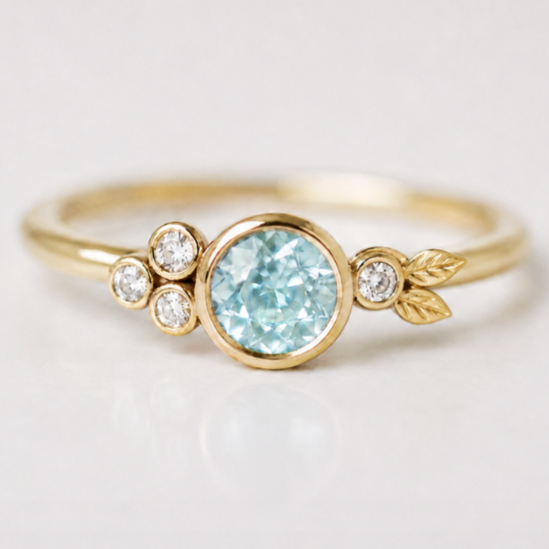 A yellow gold ring featuring a round pale green montana sapphire in the center, flanked by three small round clear stones on one side and a small leaf motif on the other, set against a clean white background.