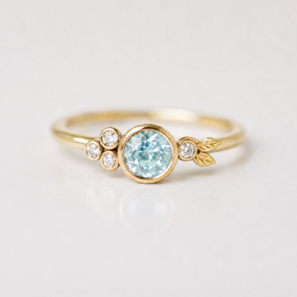A delicate yellow gold ring featuring a central round-cut light green montana sapphire, flanked by small clear accent stones and a tiny leaf detail on each side, set against a clean white background.