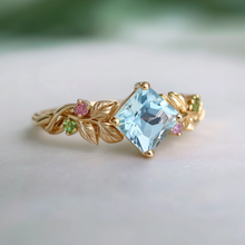 Load image into Gallery viewer, Gold ring with a blue gemstone and small pink and green stones on a light background
