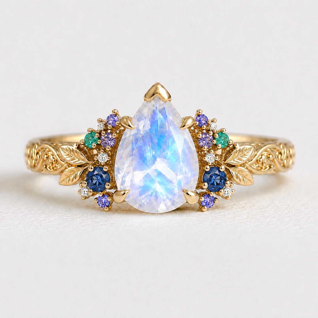 A gold ring featuring a large pear-cut iridescent moonstone center stone, accented with small blue, purple, and green gemstones and leaf-shaped details on a light gray background.